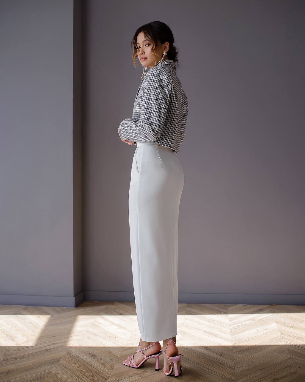 Woman wearing a checkered blouse and white pants standing in a room with a gray wall and wooden floor.