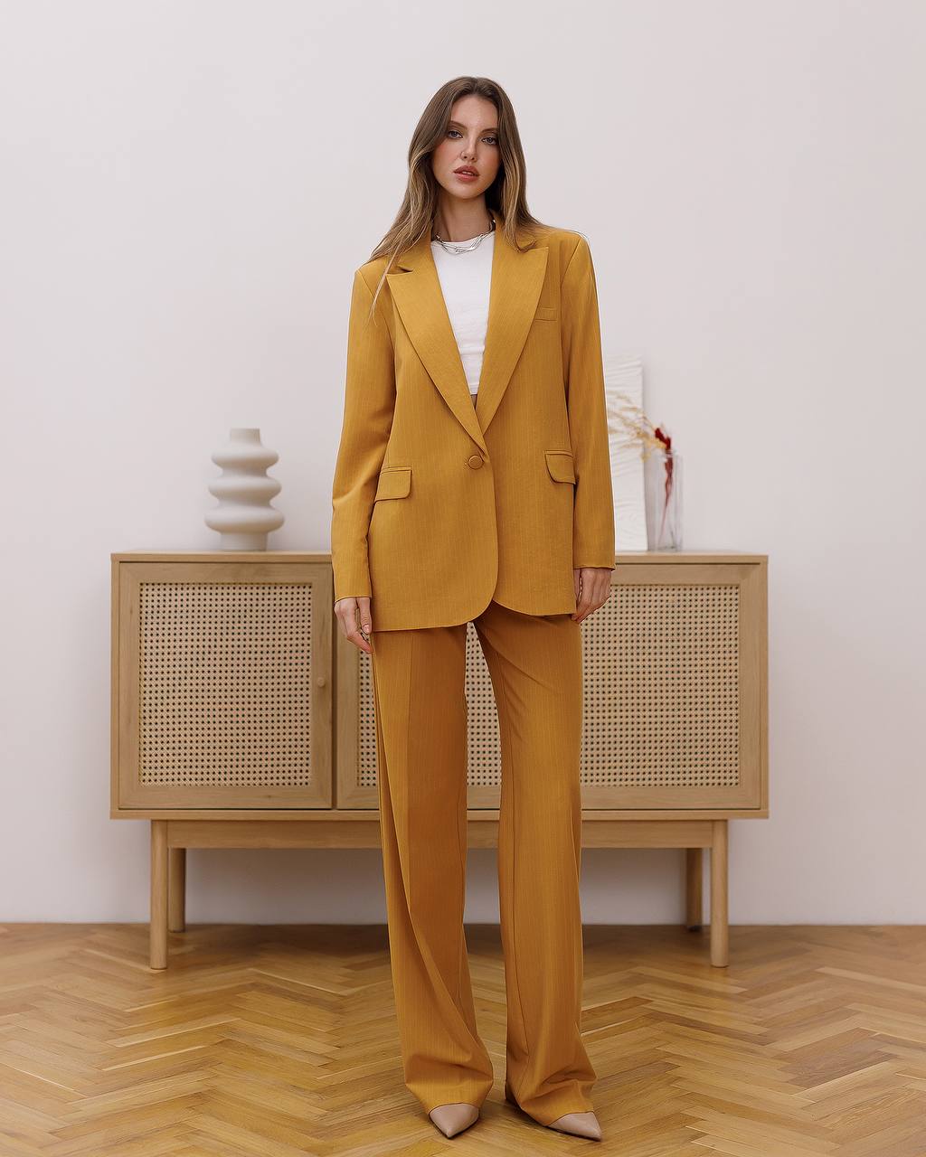 Woman wearing a mustard yellow suit standing in a room with wooden flooring and a light-colored wall.
