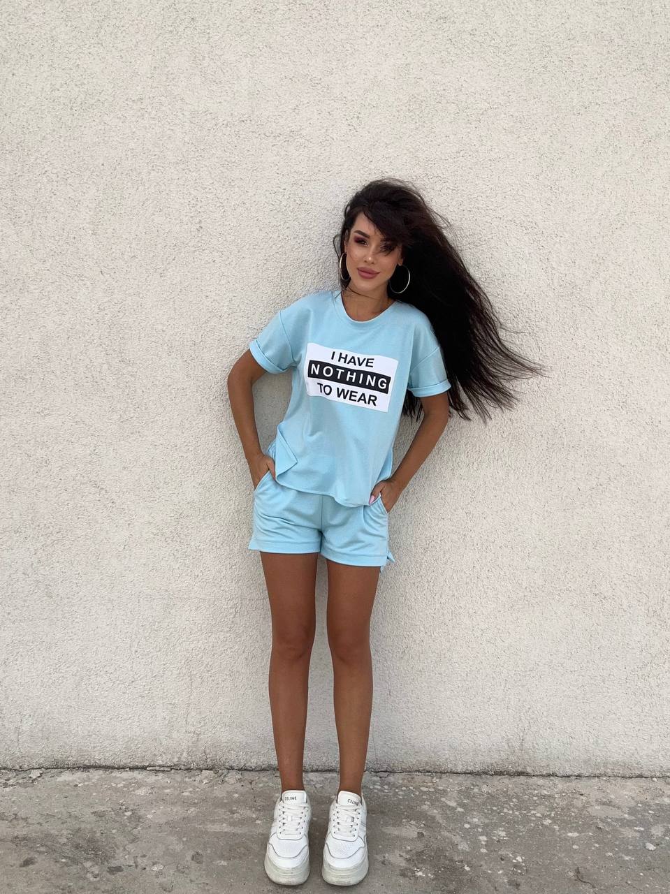 Woman wearing a light blue t-shirt and shorts with text, standing against a plain wall.