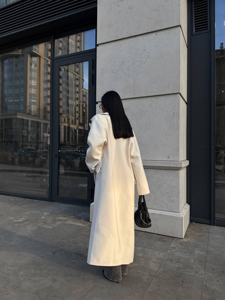 Person wearing a long white coat standing against a modern building exterior.