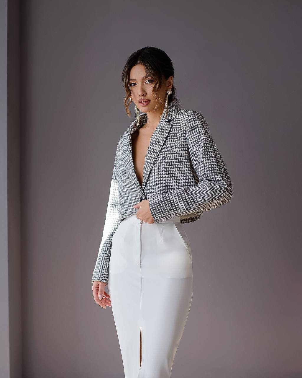 Woman wearing a checkered blazer over a deep neckline with white pants against a gray background