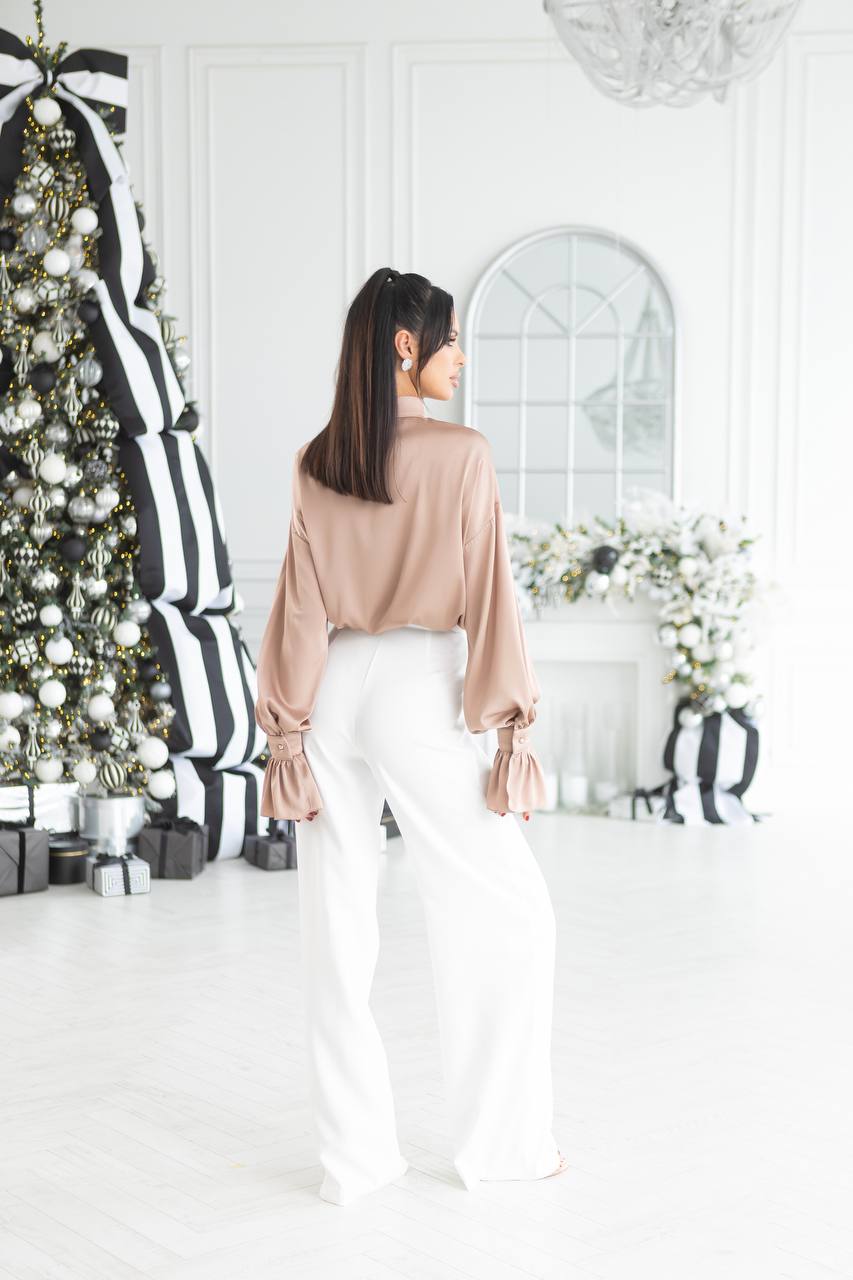 Woman in a beige blouse and white pants standing in a decorated room with Christmas trees and gift boxes.