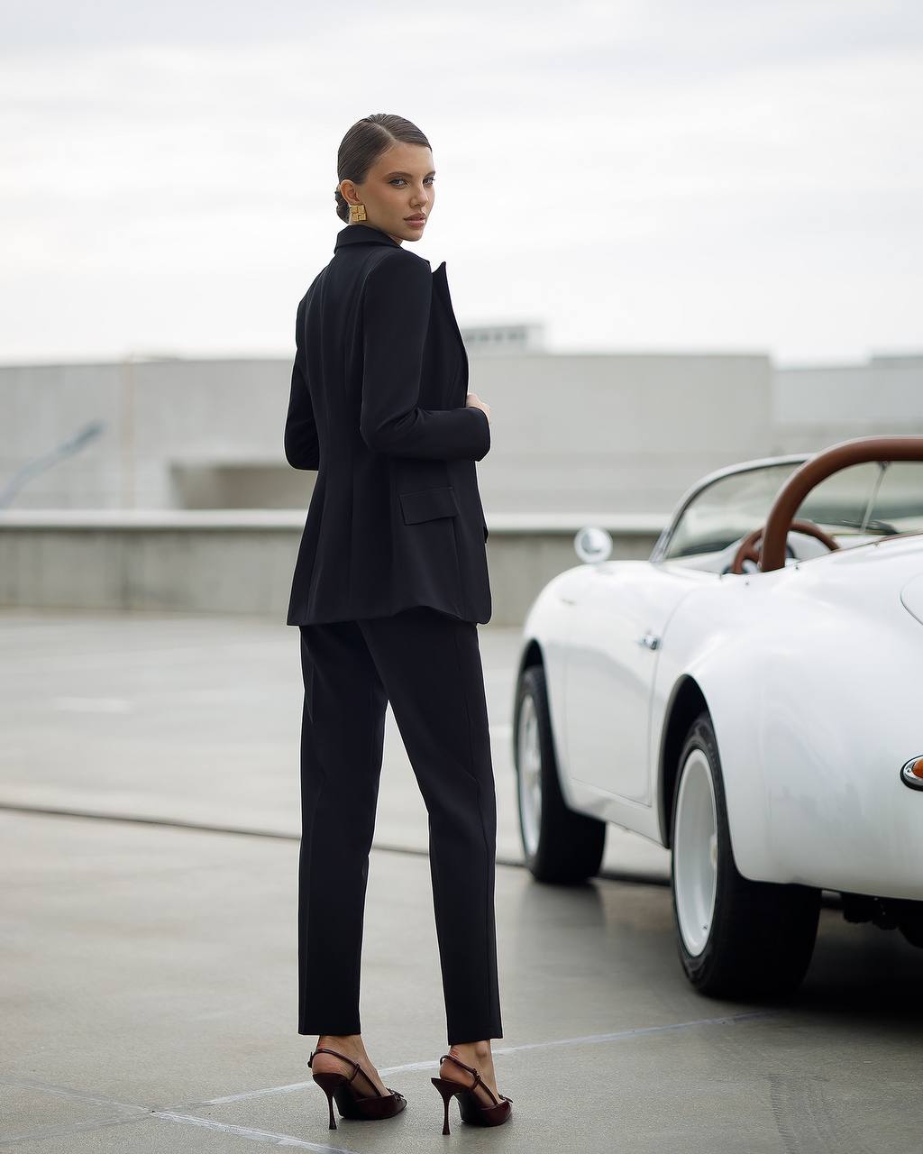 Woman in a black suit standing next to a vintage white car on a rooftop.