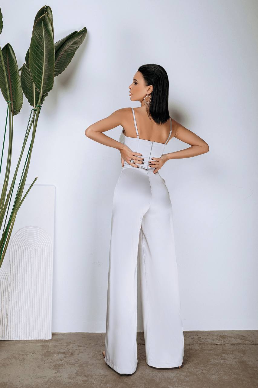 Woman wearing a white jumpsuit standing next to a plant against a plain wall.