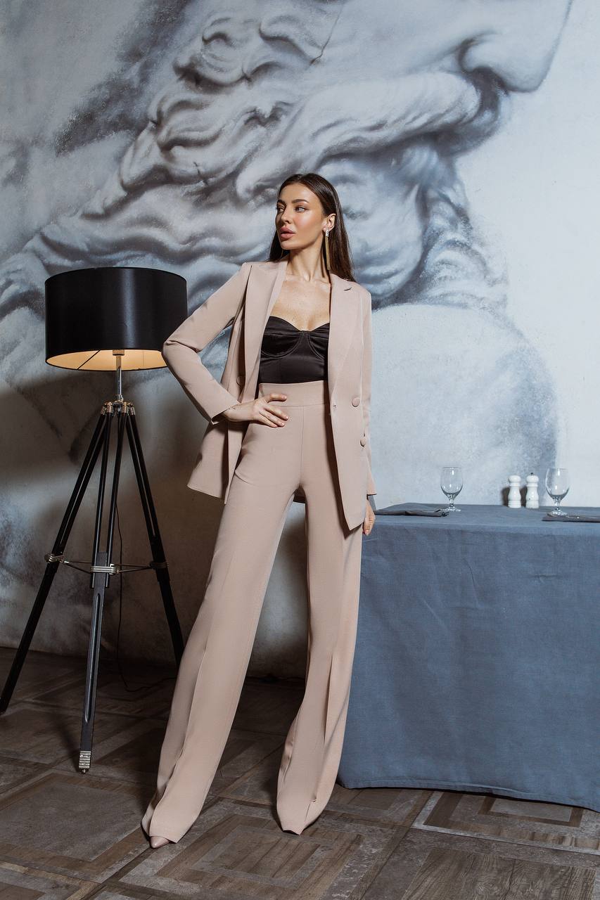 Woman in a beige suit standing in a room with a lamp and table in the background