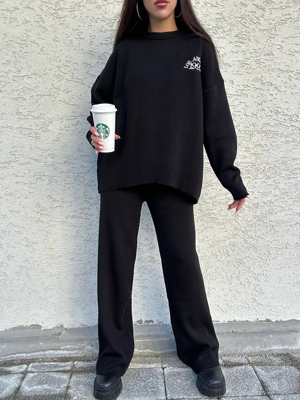 Person wearing a black outfit with a logo, holding a Starbucks cup against a light gray wall.