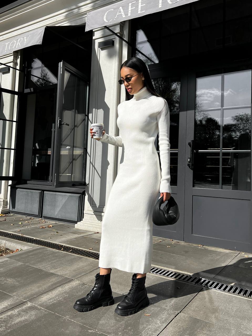 Woman in a white dress standing outside a cafe holding a coffee cup.