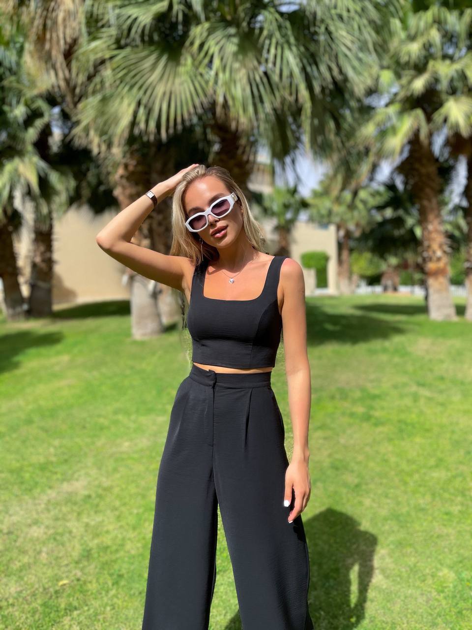 Woman in black outfit with sunglasses standing in a grassy area with palm trees