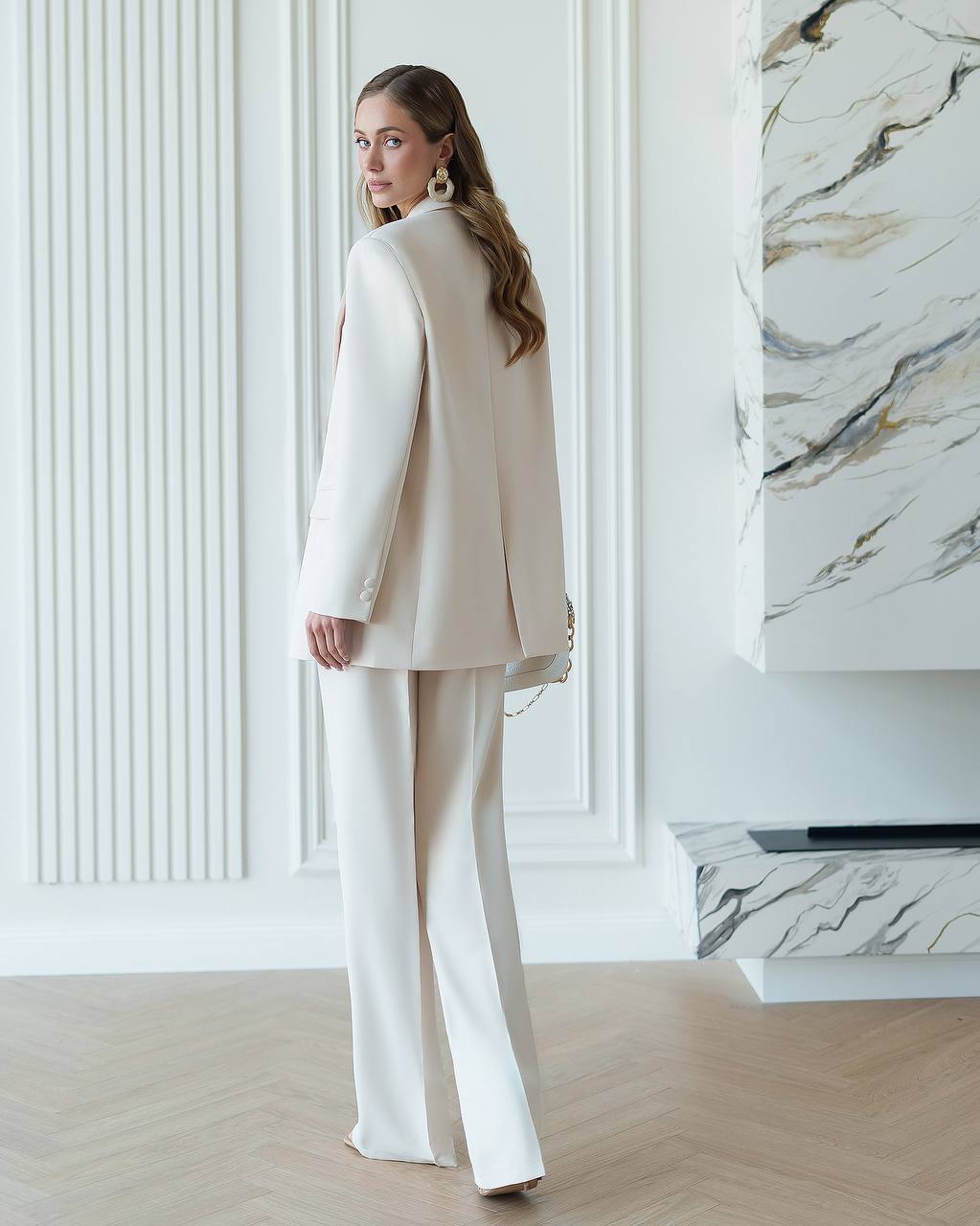 Woman in a white suit standing in a stylish interior setting with marble accents.