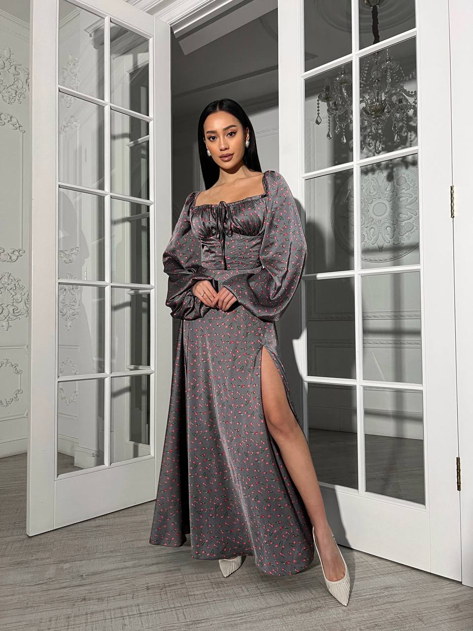 Woman in a long, dark patterned dress standing in a room with large windows.