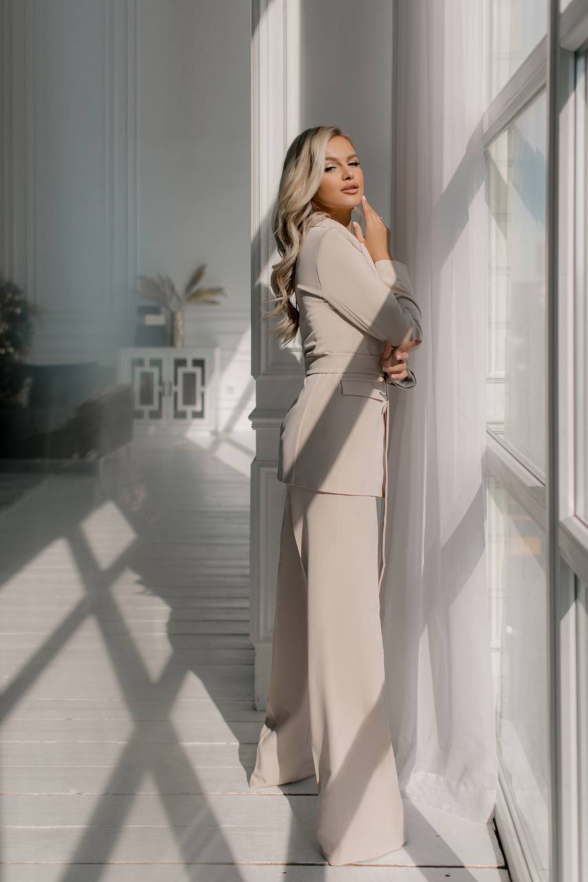 Woman in a beige suit standing in a sunlit room with shadows on the wall.
