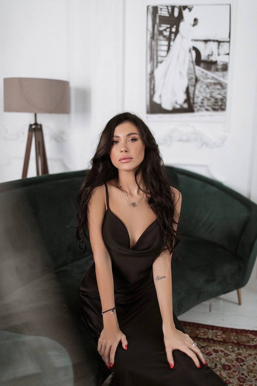 Woman in a black dress sitting on a green couch in a room with a lamp and framed picture on the wall.