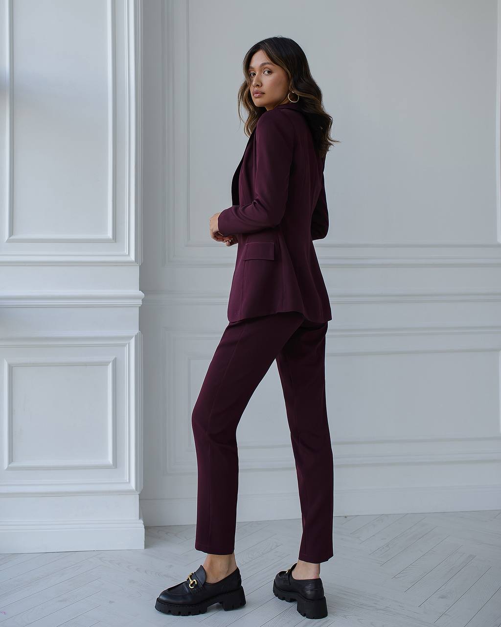 Woman wearing a burgundy suit standing against a white paneled wall.