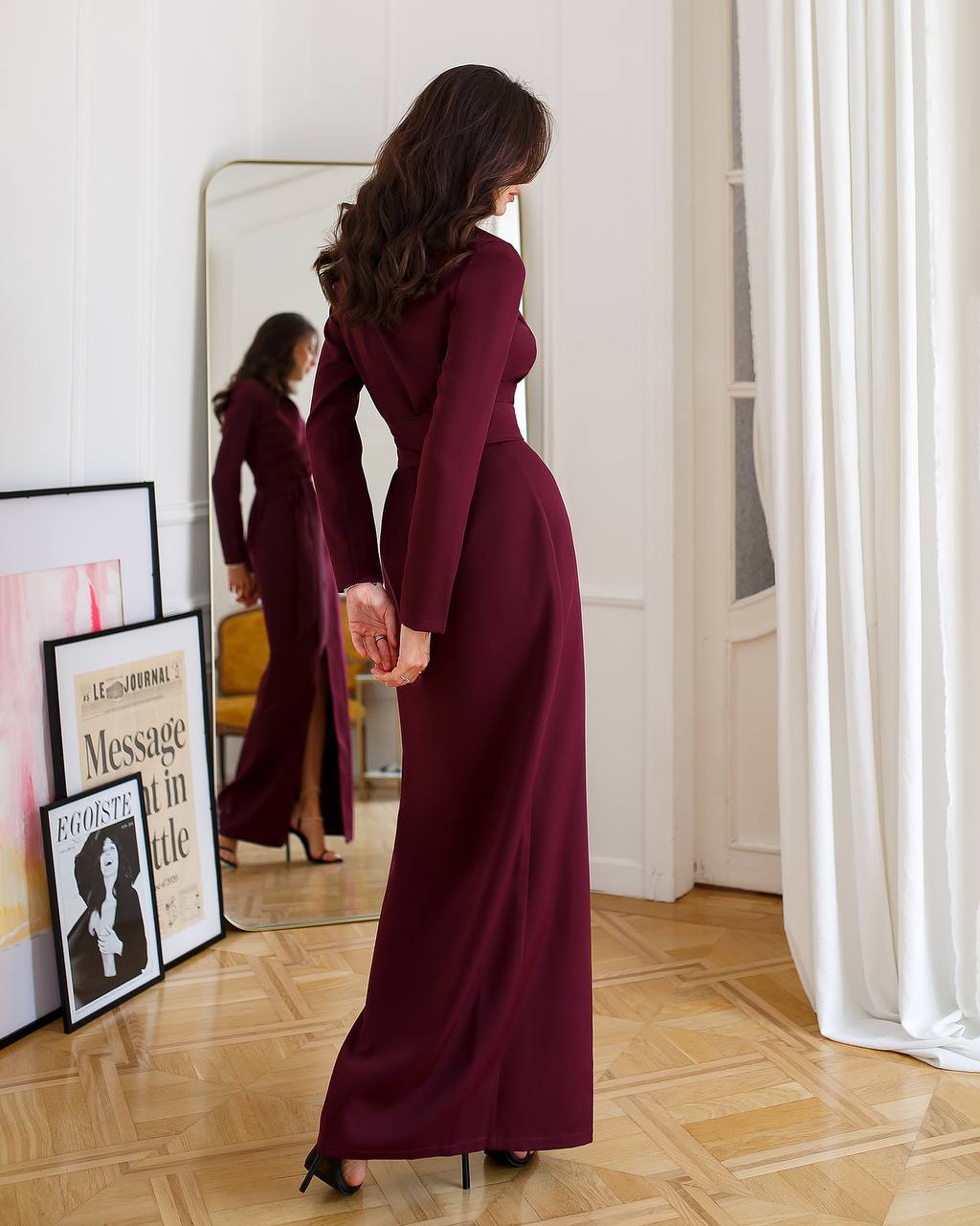 Woman in a burgundy dress standing in a room with a mirror and framed pictures on the wall.