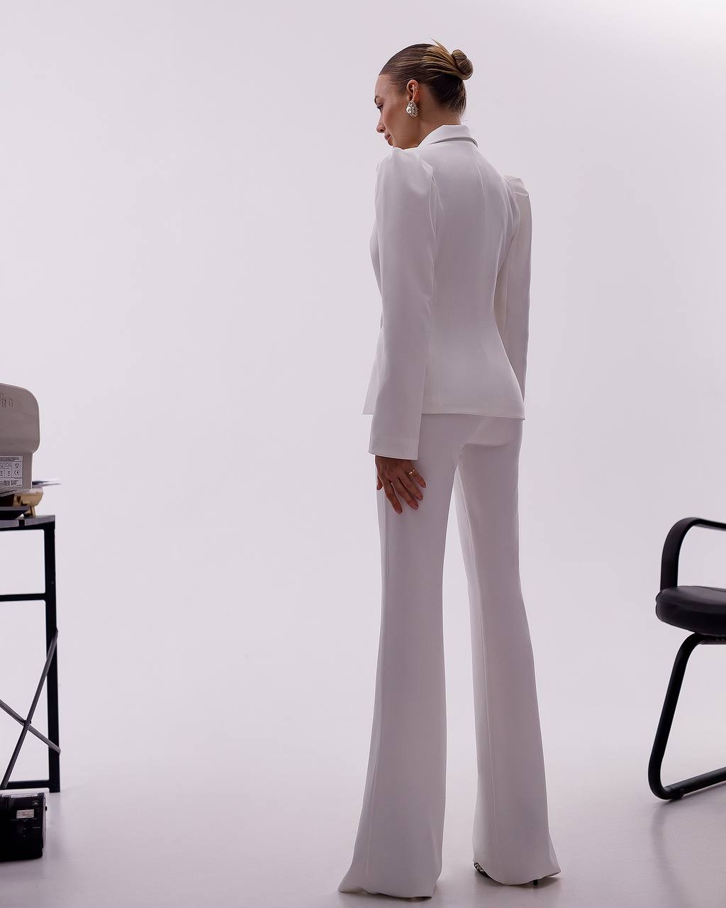 Person wearing a white suit standing in a minimalistic room with a chair and table.