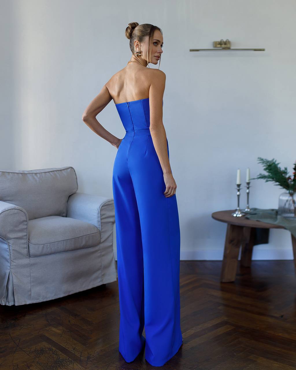 Woman in a blue strapless jumpsuit standing in a room with a gray armchair and wooden table.