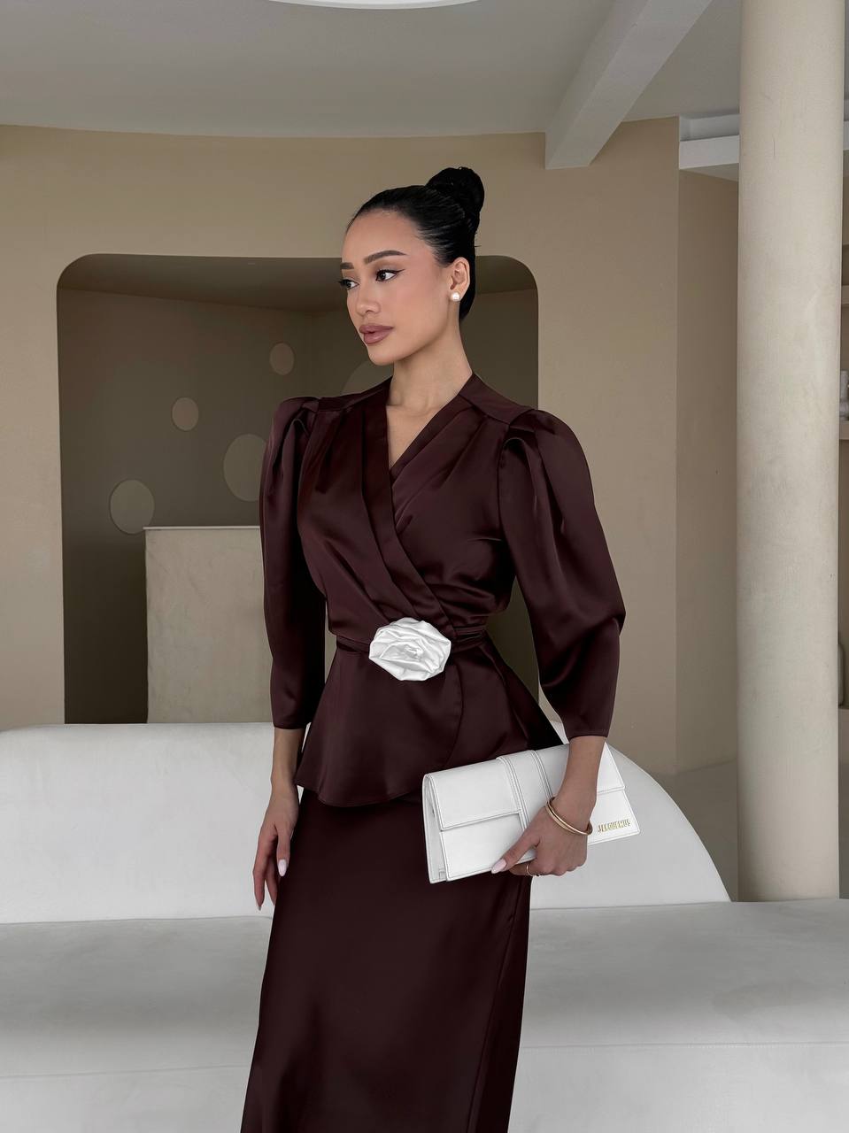 Woman in a dark brown outfit holding a white clutch in an indoor setting