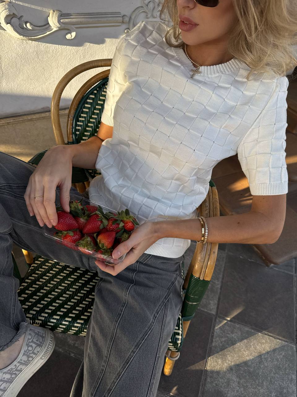 Person sitting outdoors holding a plate of strawberries, wearing a white textured top and gray pants.