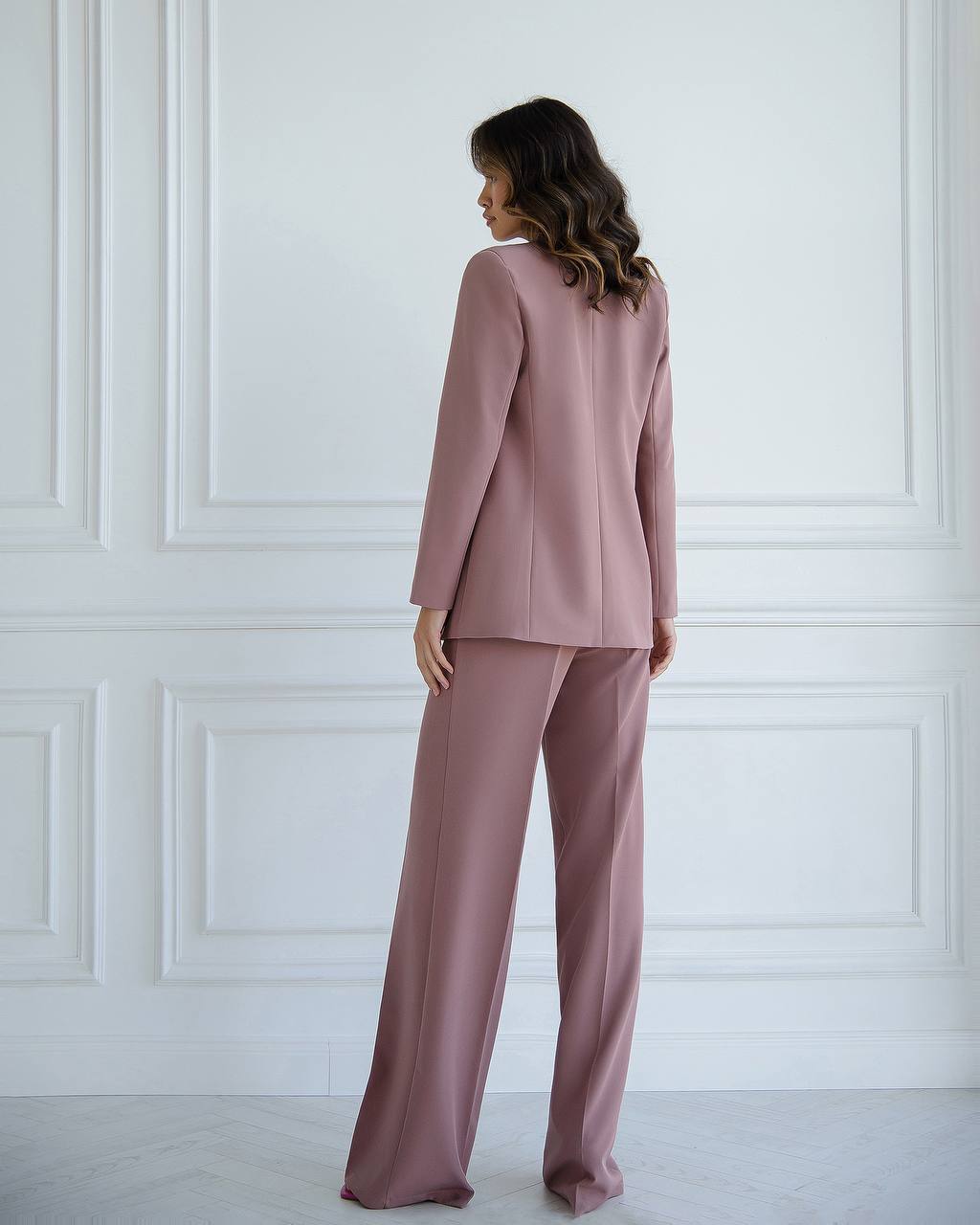 Woman wearing a pink suit standing against a white paneled wall.