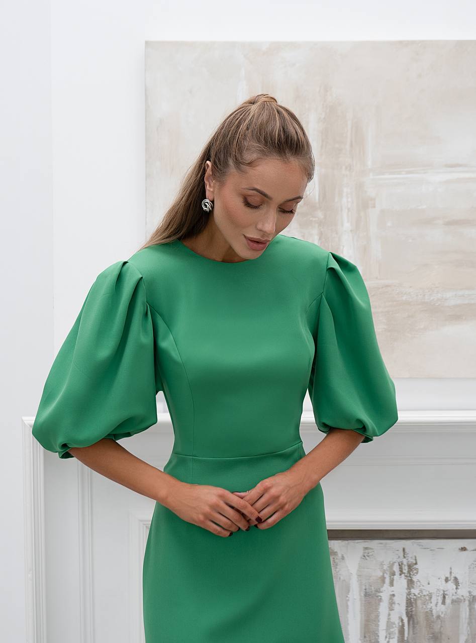 Woman wearing a green dress with puffed sleeves against a light-colored wall.