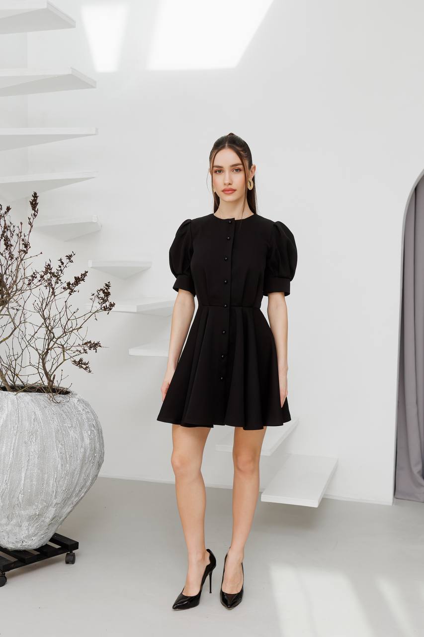 Woman wearing a black dress standing in a minimalistic room with white walls and a plant.
