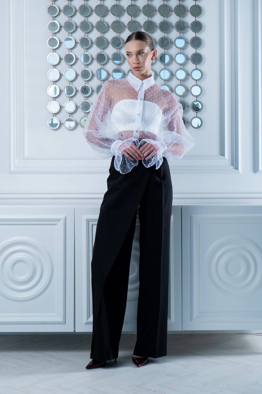 Woman wearing a sheer white blouse and black pants against a decorative wall.