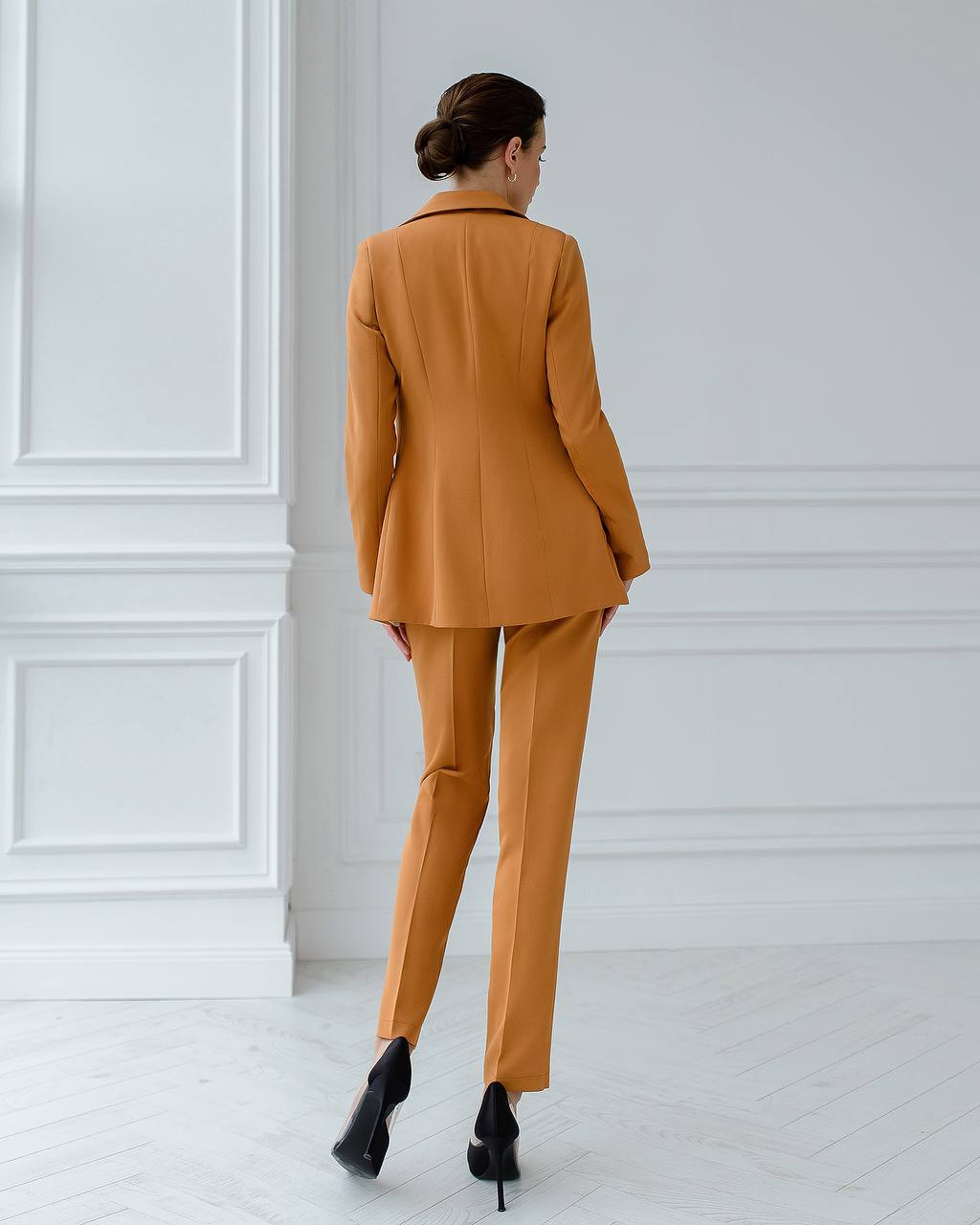 Woman wearing a mustard yellow suit standing against a white paneled wall.