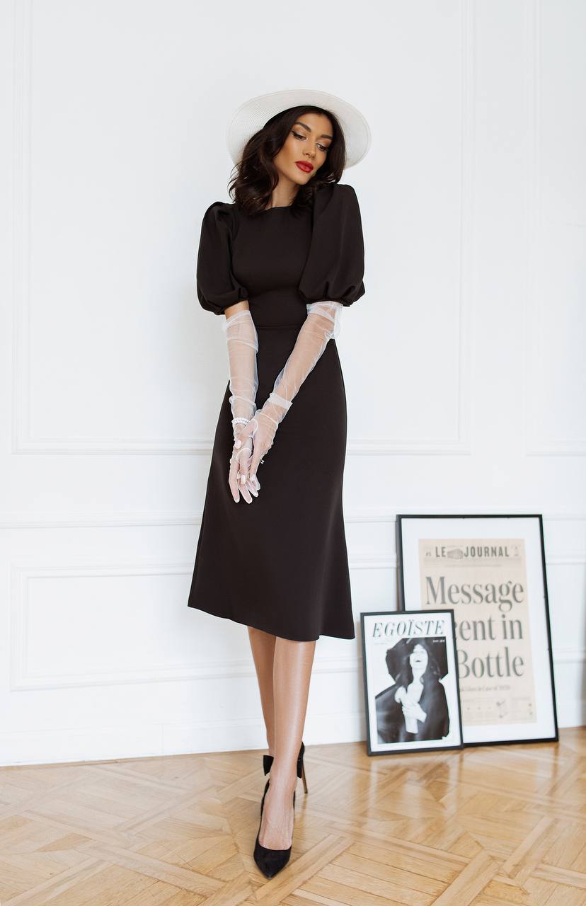 Woman in a black dress with white gloves standing in a room with framed pictures on the wall.