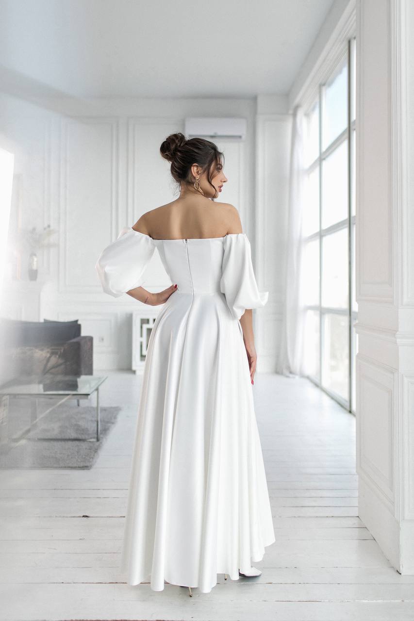 Woman in a white off-shoulder dress standing in a bright, minimalistic room.