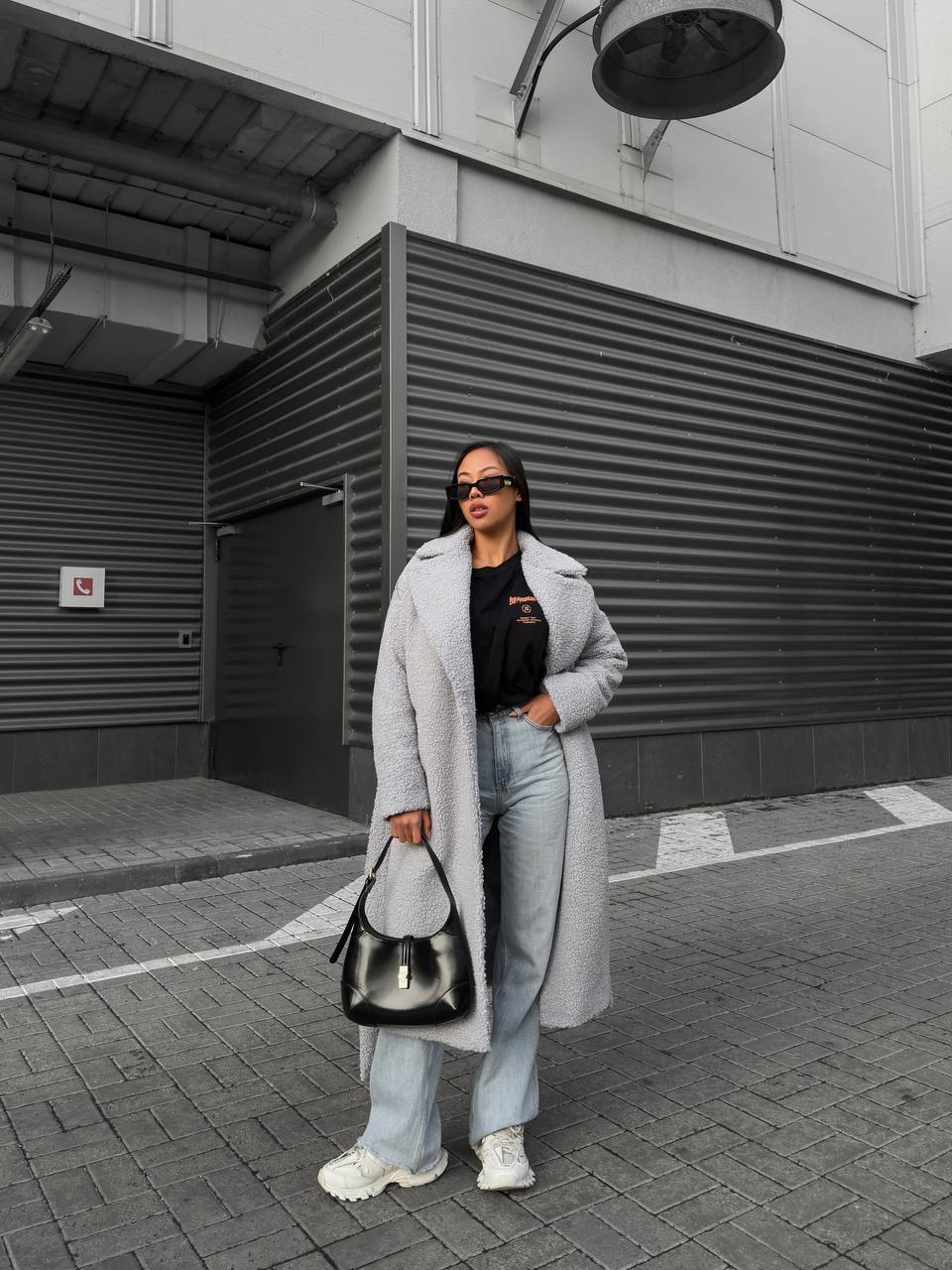 Person wearing a long gray coat and light blue jeans standing on a street.