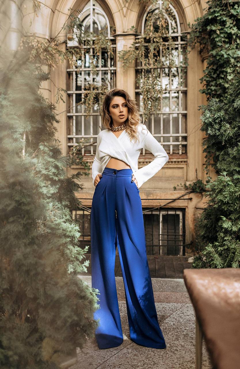 Woman in a white top and blue pants standing in an outdoor setting with architectural elements and greenery.