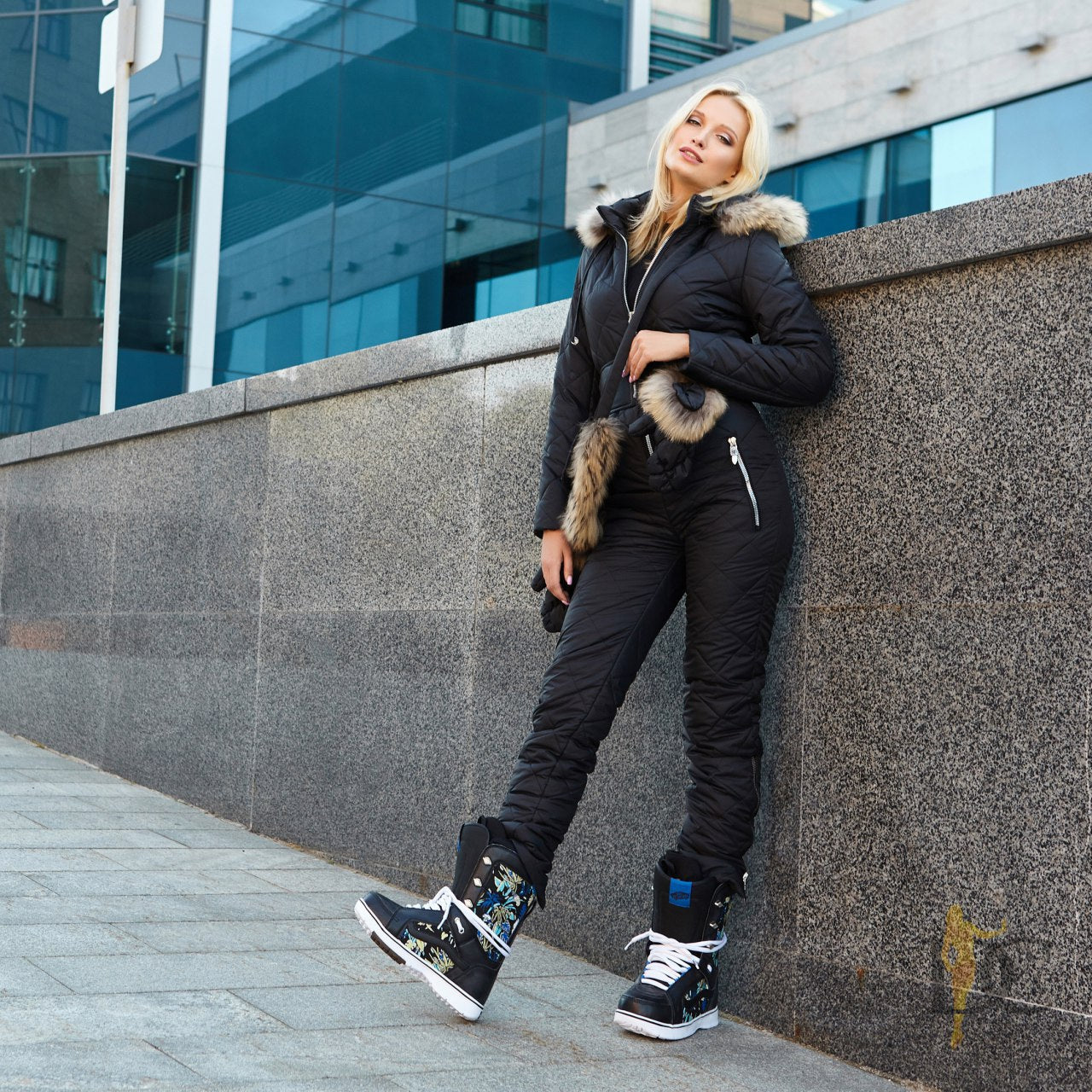 Woman in black winter outfit with fur-lined hood leaning against a wall in an urban setting.