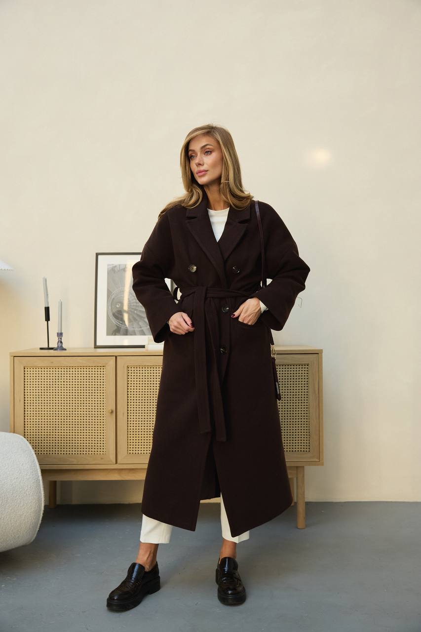 Woman wearing a long dark coat in a room with a wooden cabinet and lamp.