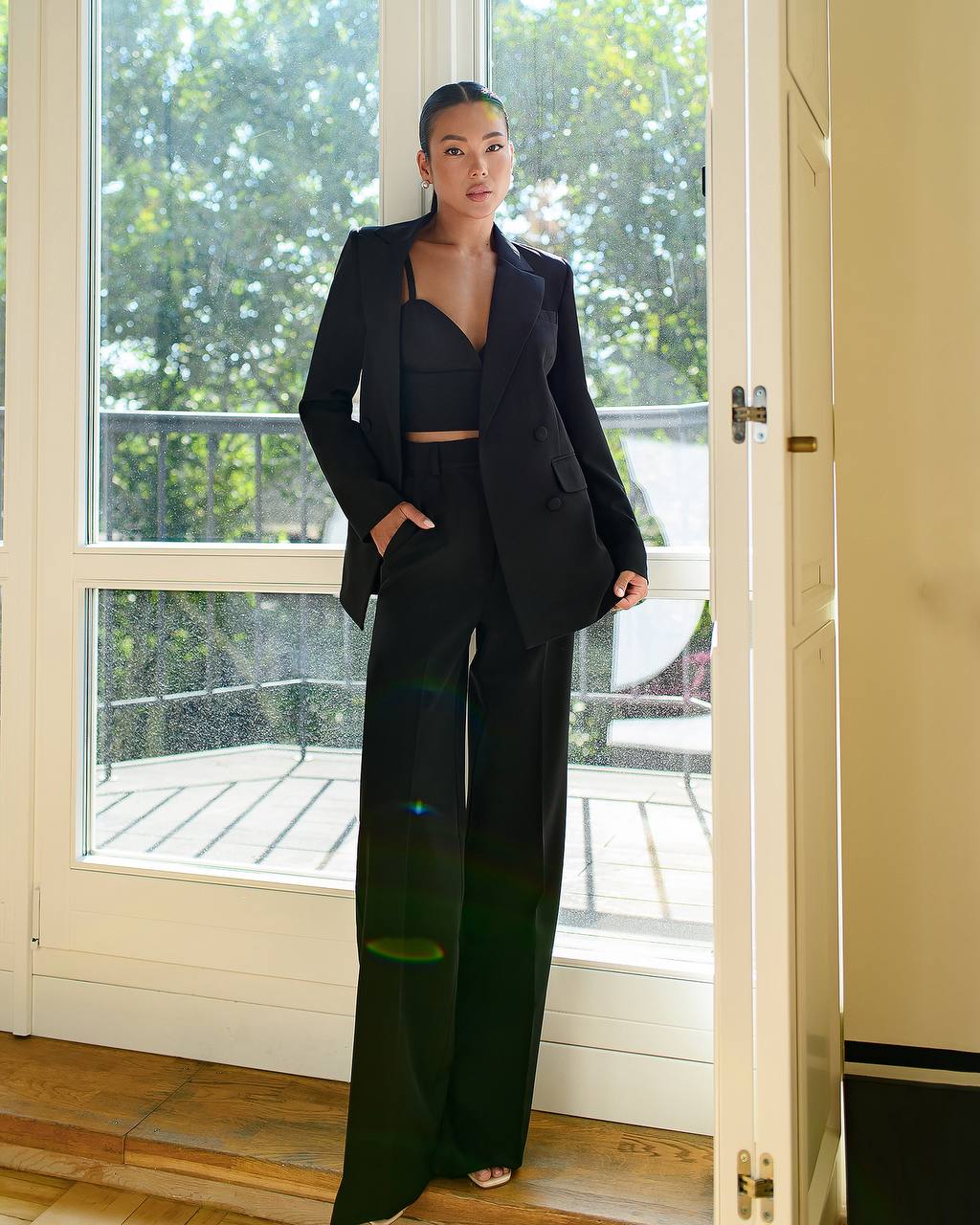 Woman in a black suit standing in a room with a large window showing greenery outside.
