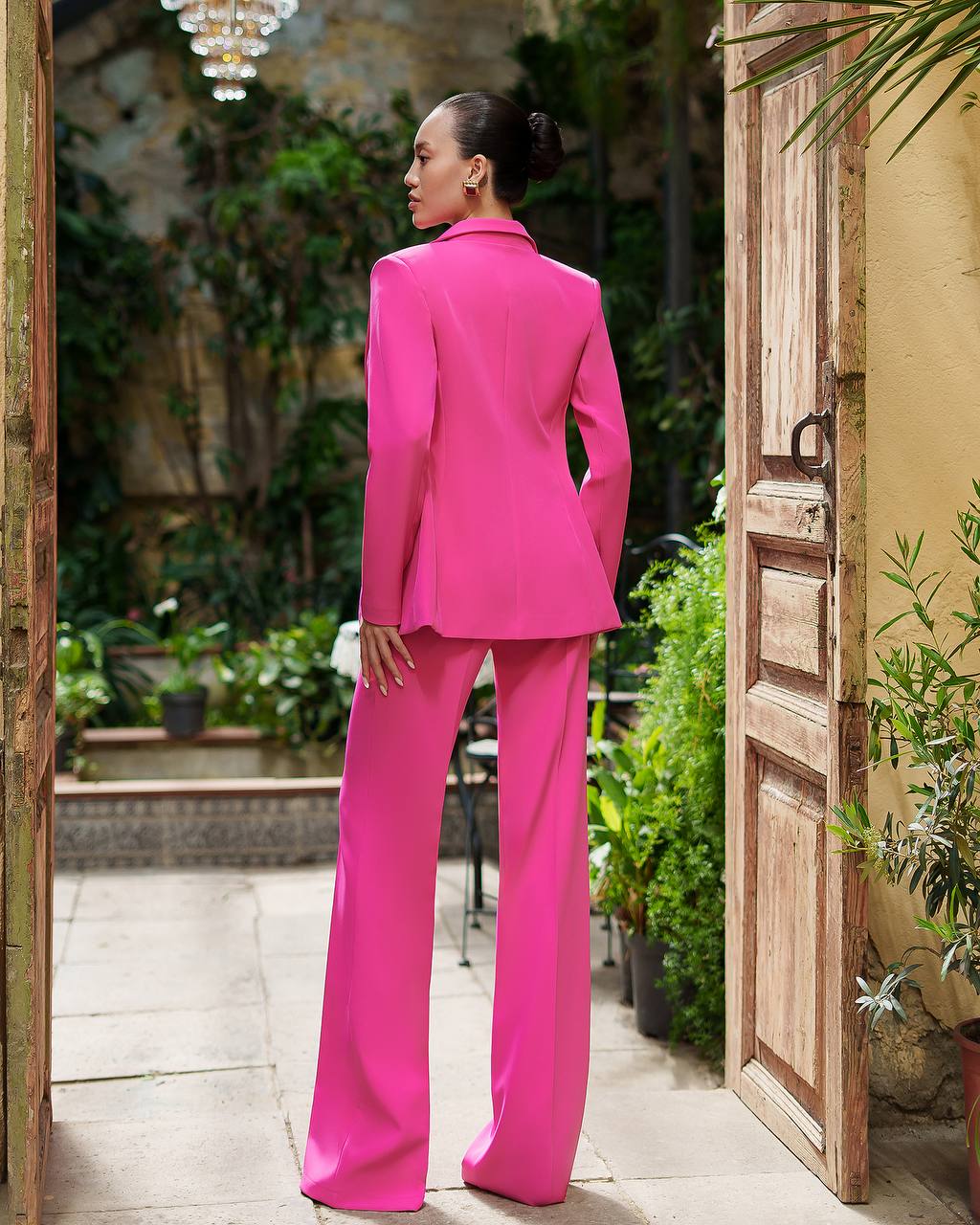 Person wearing a bright pink suit standing in an outdoor setting with plants and a wooden door.