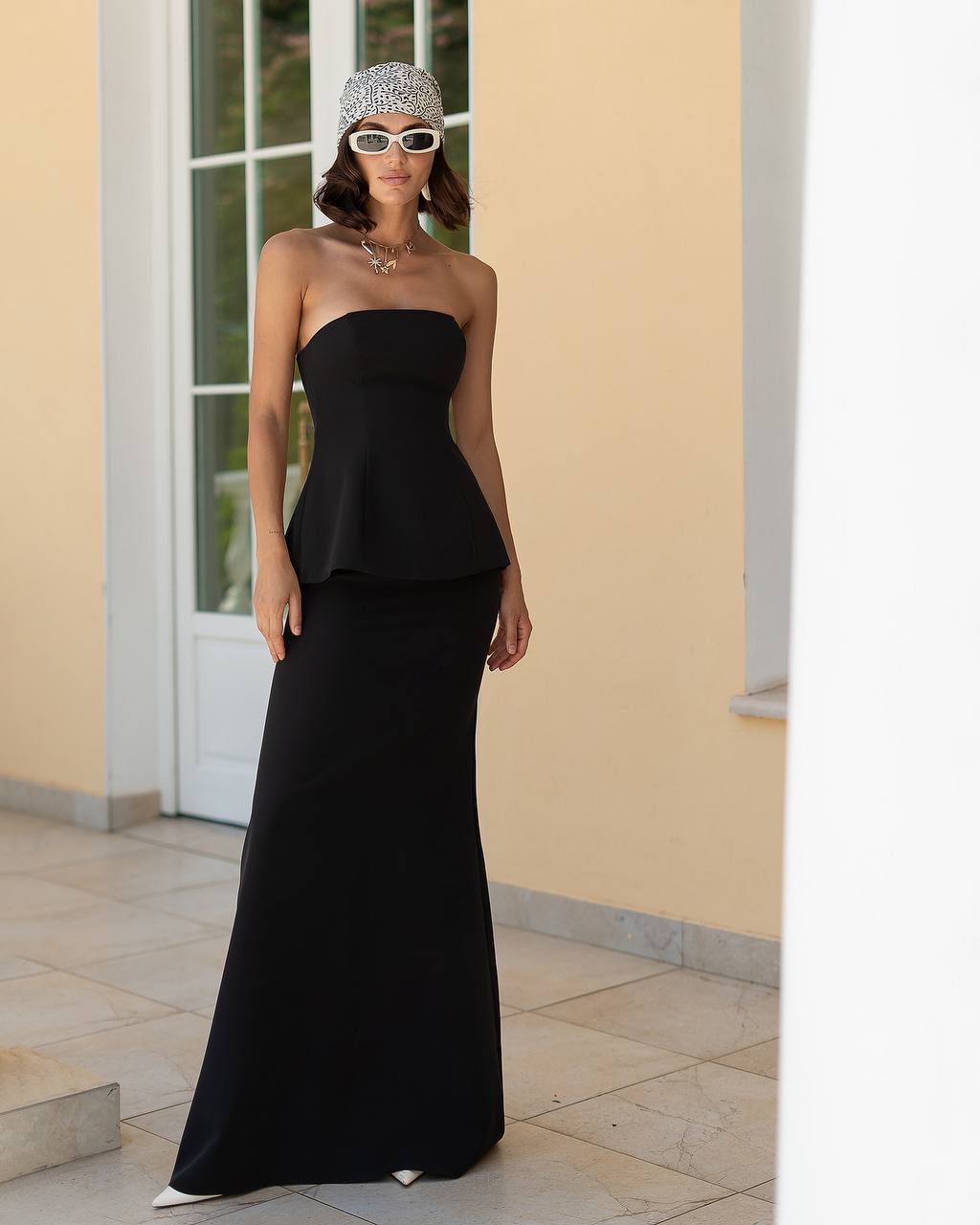 Woman wearing a black strapless dress and sunglasses in front of a beige wall.