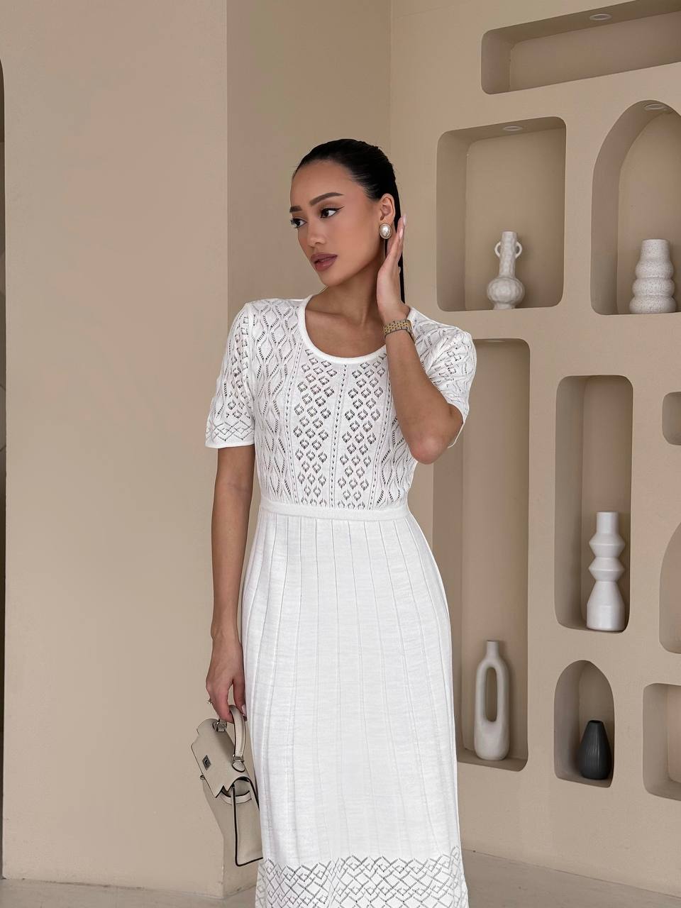 Woman wearing a white lace dress standing in front of a decorative wall with shelves.