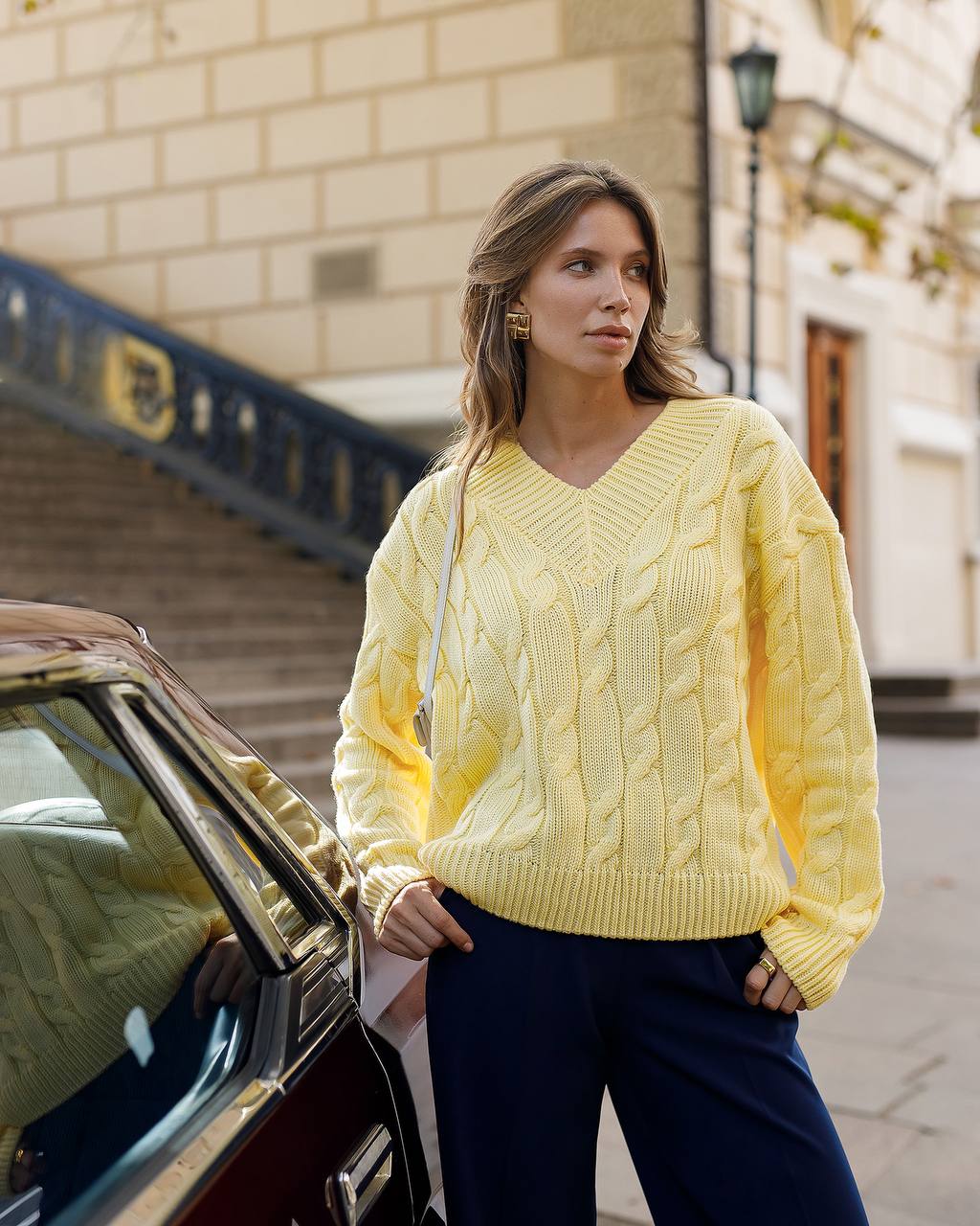 Woman wearing a yellow cable knit sweater standing next to a car on a city street.