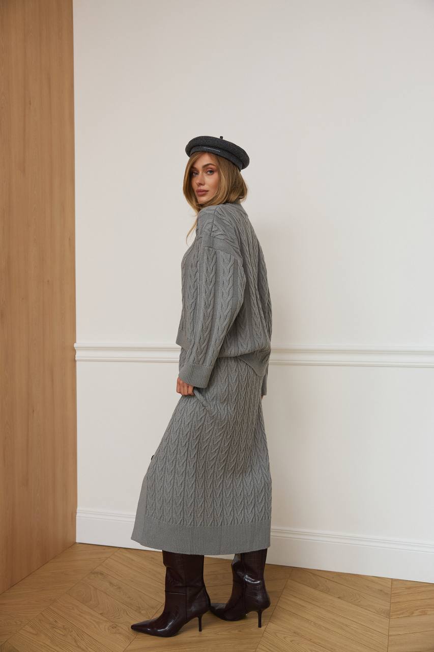 Woman wearing a gray cable knit dress with a matching cardigan and black boots, standing against a white wall.
