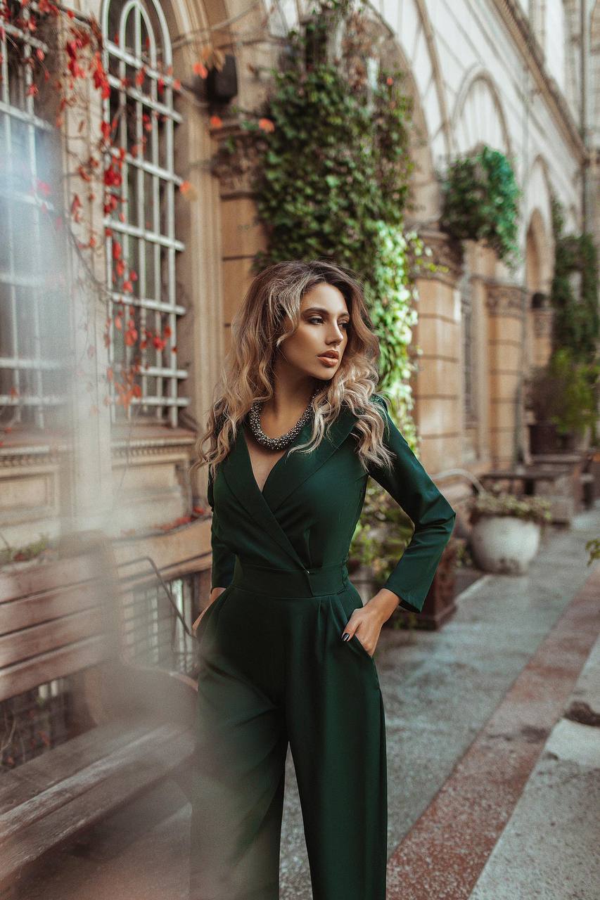 Woman in a green jumpsuit standing in an outdoor setting with architectural elements and plants.