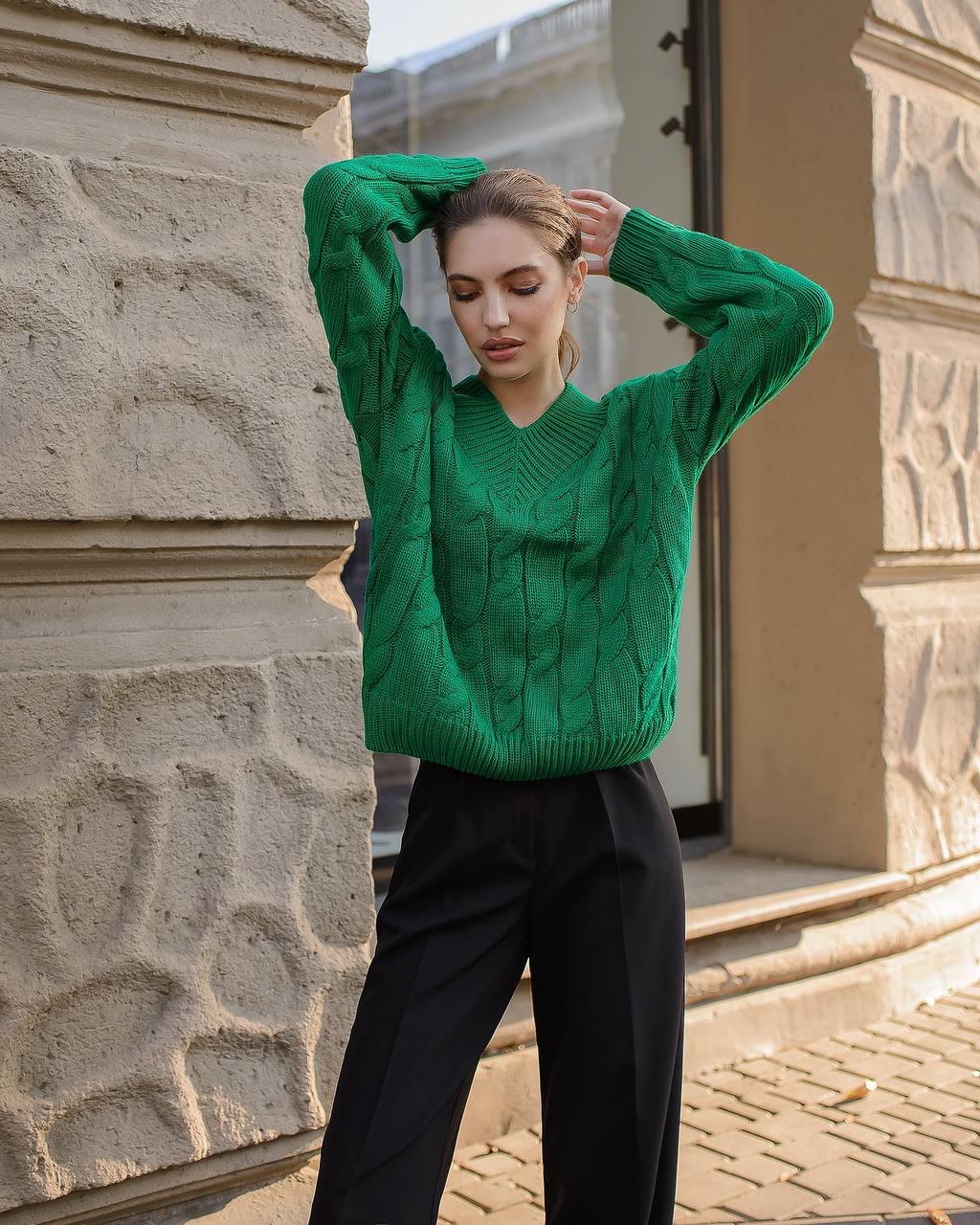Woman wearing a green sweater and black pants standing in front of a building.