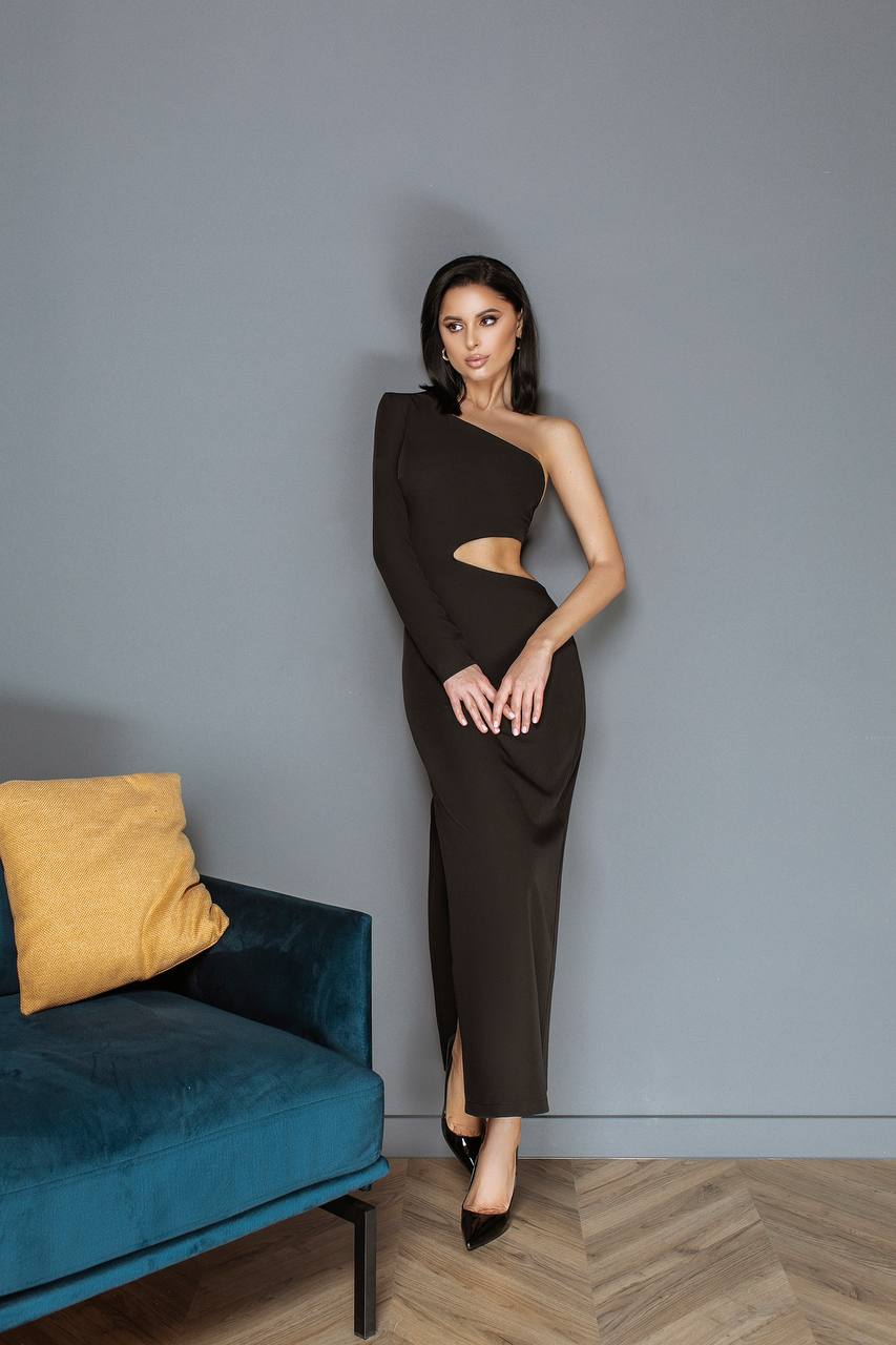 Woman in a black one-shoulder dress standing against a gray wall with a blue couch and yellow pillow.