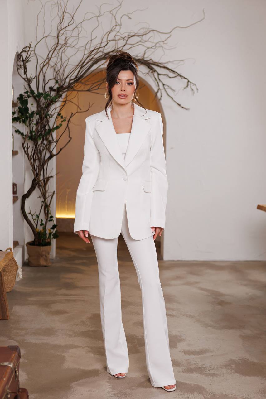 Woman in a white suit standing in a room with a plant and wooden furniture.
