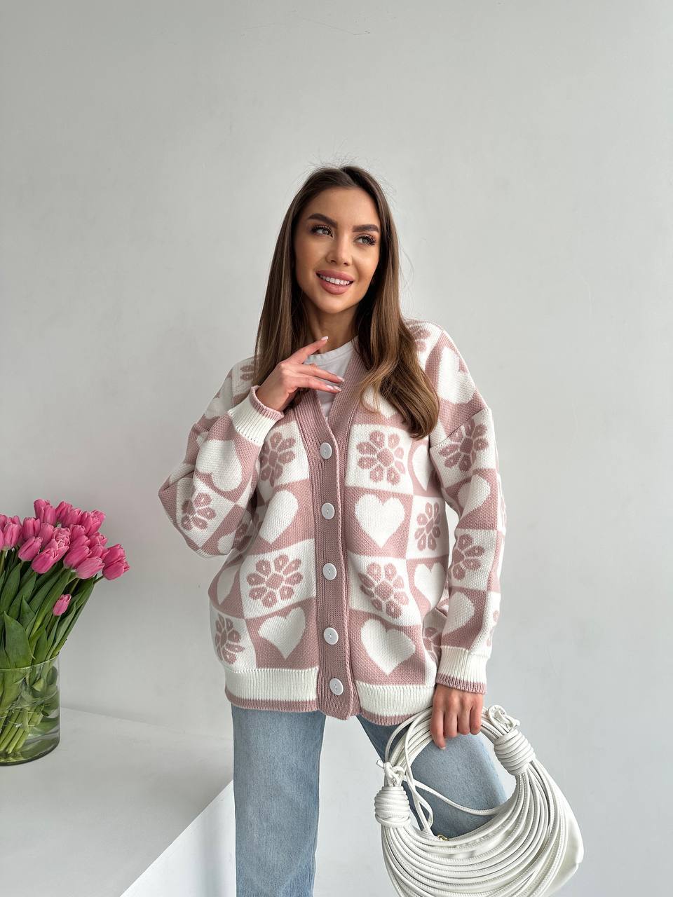 Woman wearing a patterned cardigan with pink tulips and a white bag on a light background