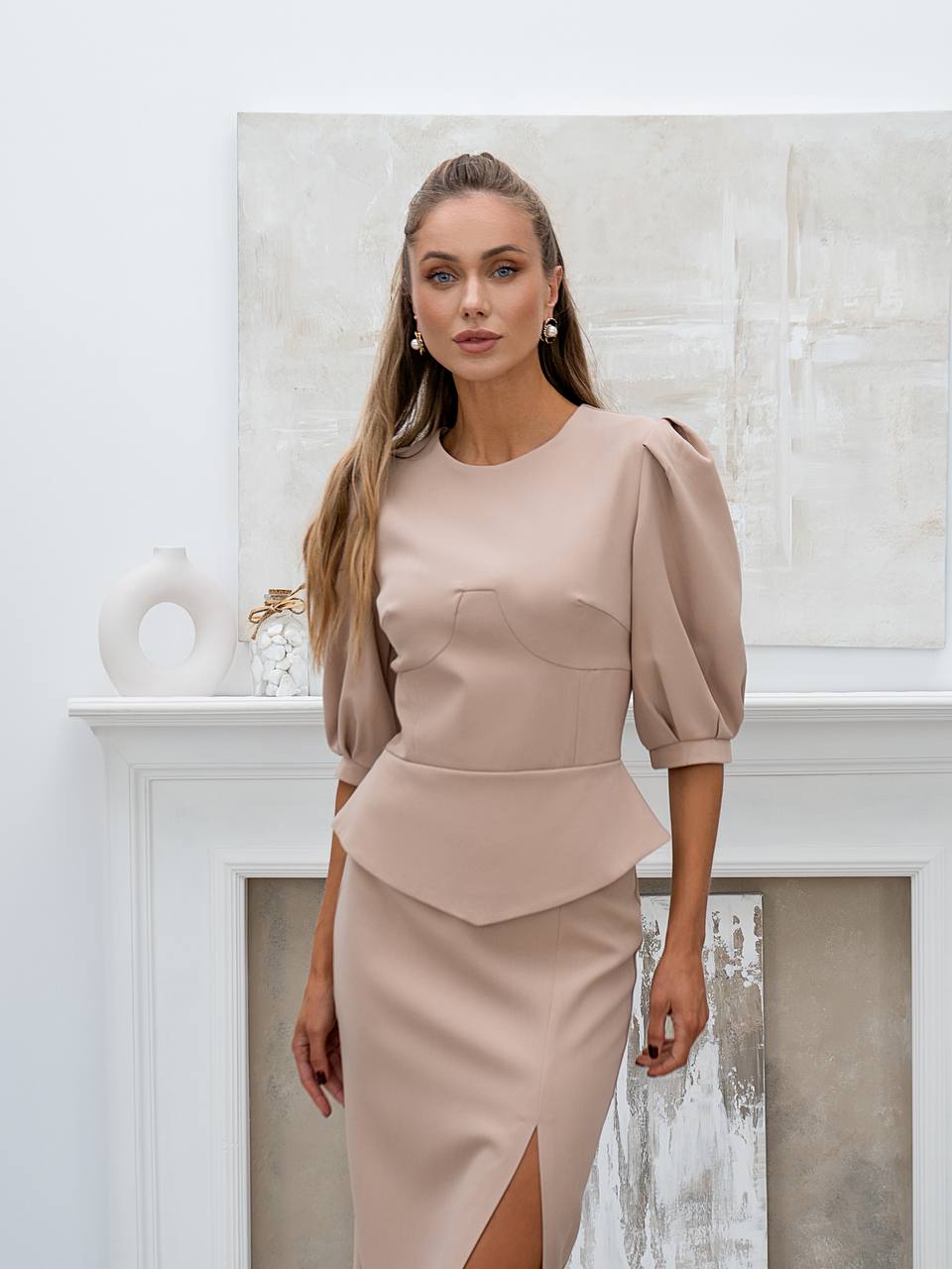Woman wearing a beige peplum top and skirt in a white room.