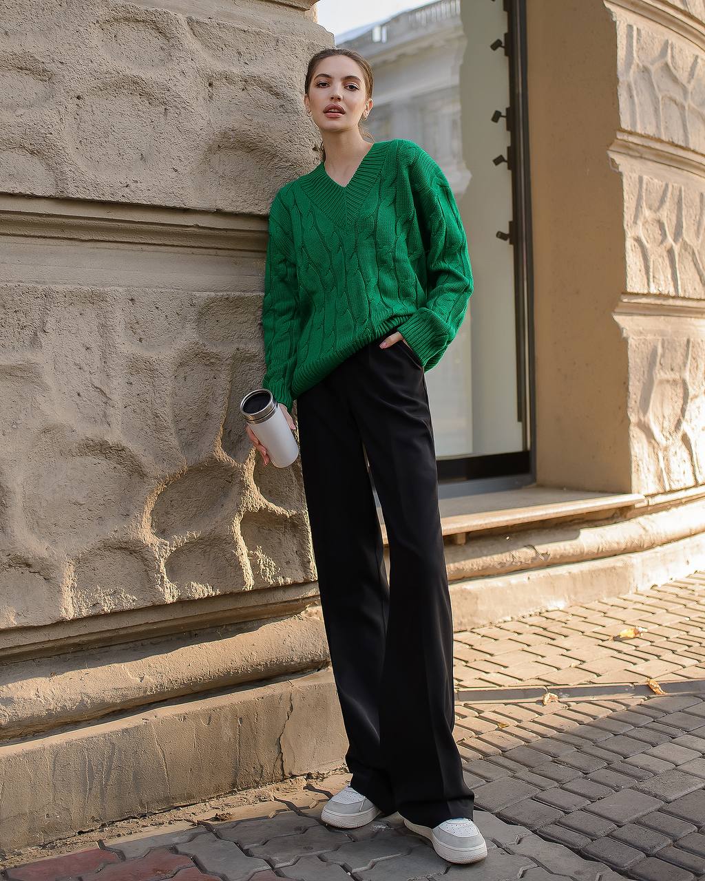 Person wearing a green sweater and black pants standing against a stone wall.