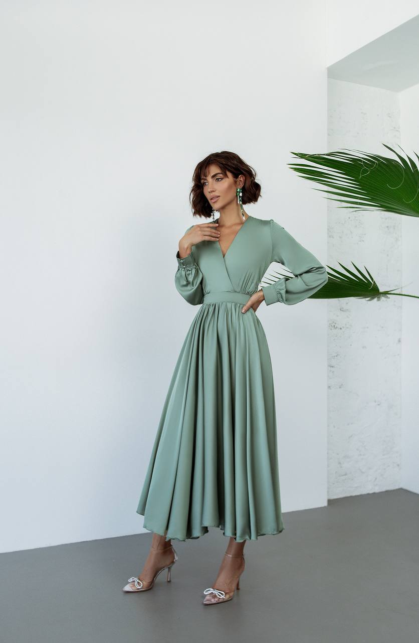 Woman wearing a green dress standing against a white wall with a plant.