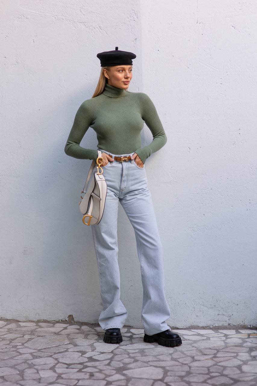 Person wearing a green turtleneck, light blue jeans, black shoes, and a black beret against a white wall.