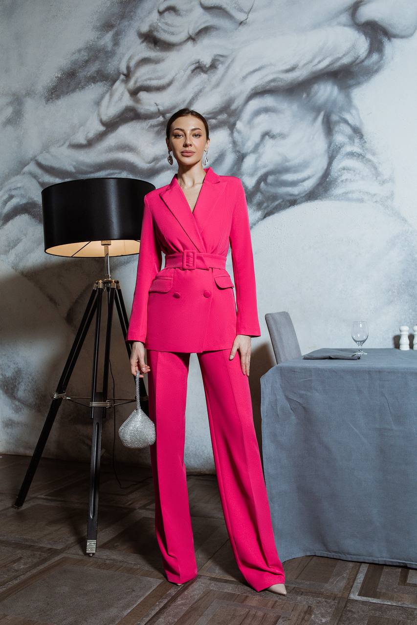 Woman in a bright pink suit standing in an elegant room with a lamp and table.
