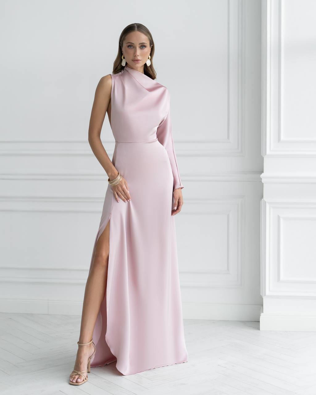 Woman wearing a pink evening gown with a high slit against a white wall.